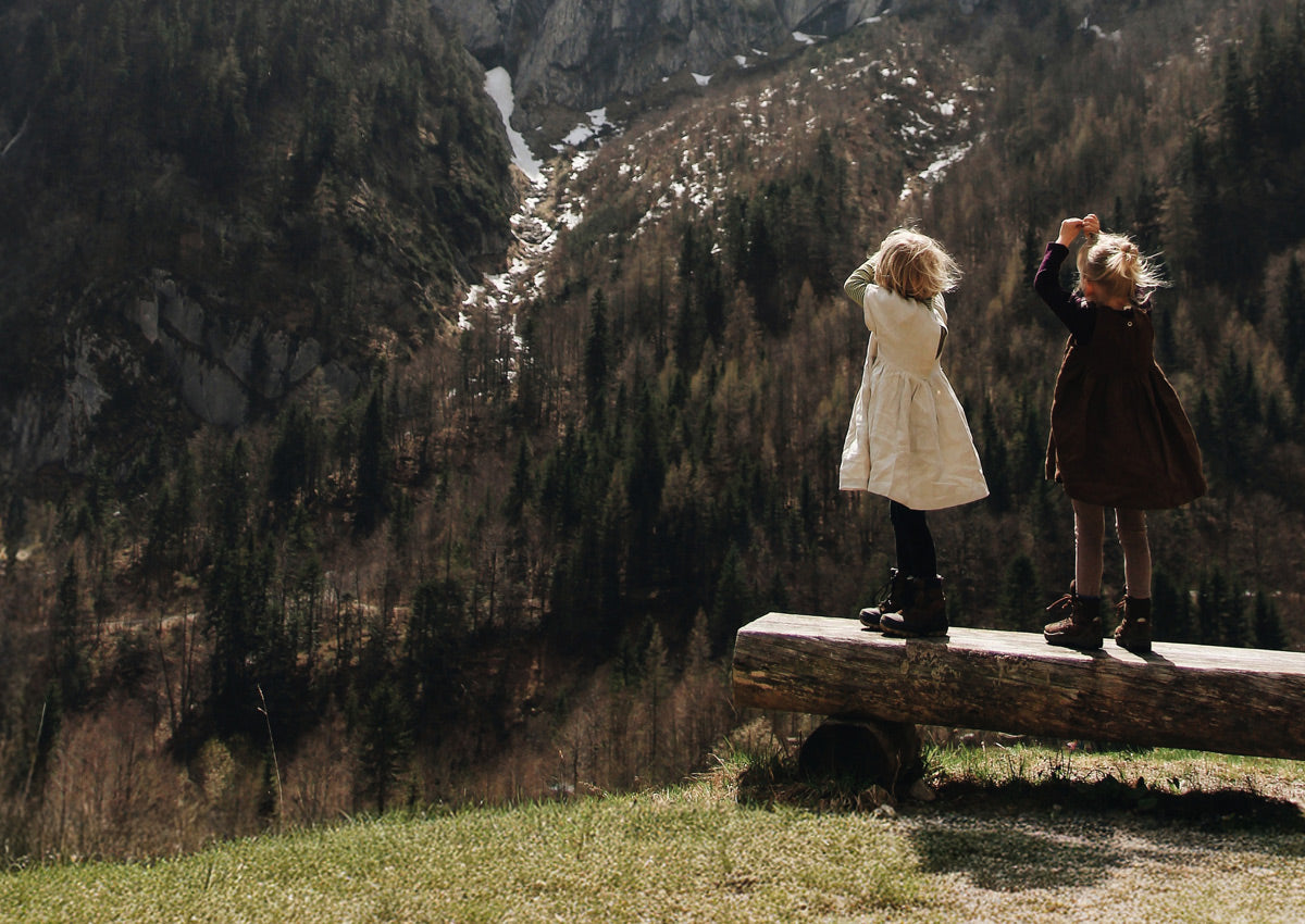 Zwei Kinder stehen auf einer Holzbank in den Bergen, ihre Kleider wehen